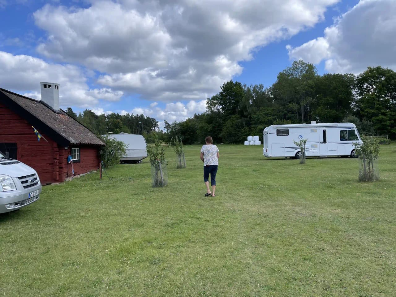 Alt-text: En grönskande hembygdsgård med en stor gräsmatta, husvagnar och husbilar parkerade omkring området. Besökarna njuter av den natursköna omgivningen.