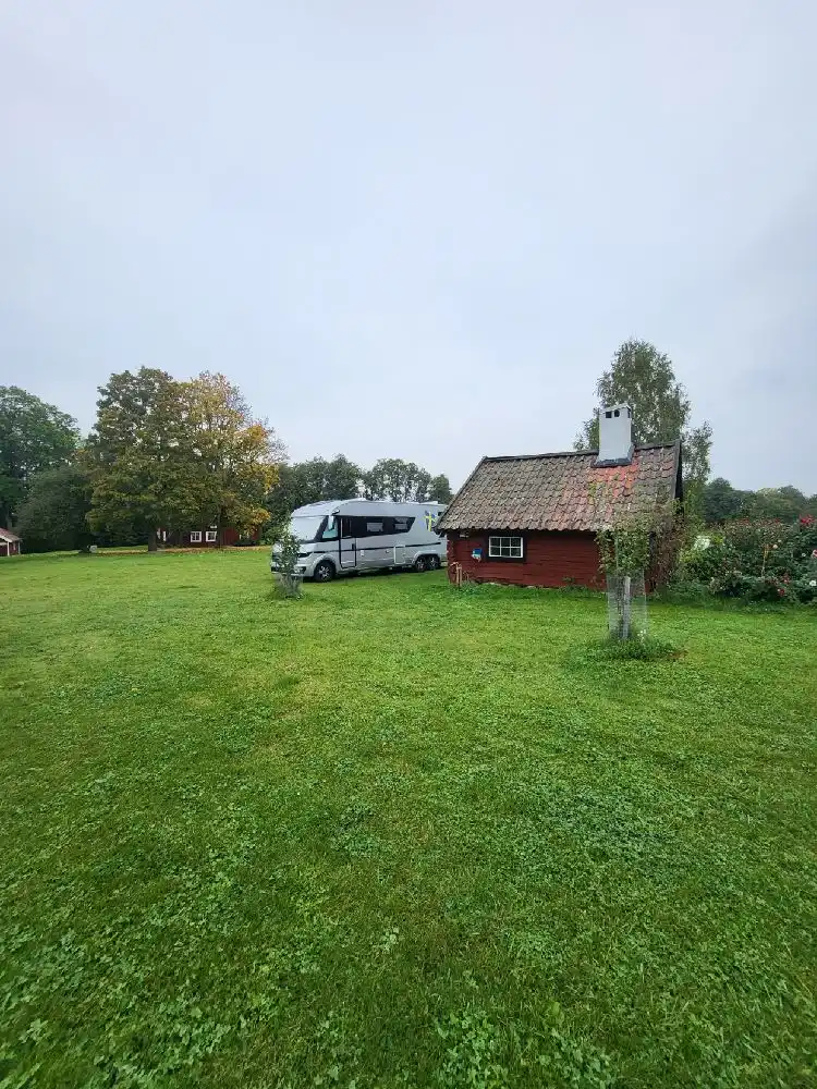 En grönskande hembygdsgård med en stuga omgiven av gräs och träd. I förgrunden syns en familjebil parkerad på gårdsplanen.