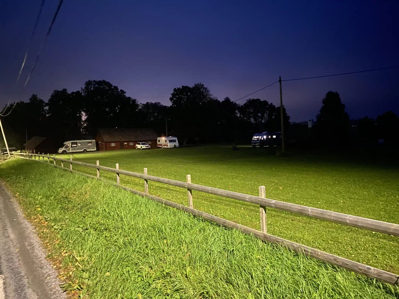 En naturskön vy över Kvarngården hembygdsgård visar en gräsbevuxen gård omgiven av ett klassiskt trästaket. Bilden är tagen i skymning, vilket skapar en mjuk linsflare som förhöjer den lantliga atmosfären.