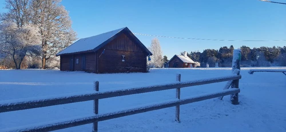 Bild av en pittoresk hembygdsgård i vinterlandskap med ett snötäckt tak. Den omgivande naturen med trädgrenar täckta av snö förstärker den lugna atmosfären.