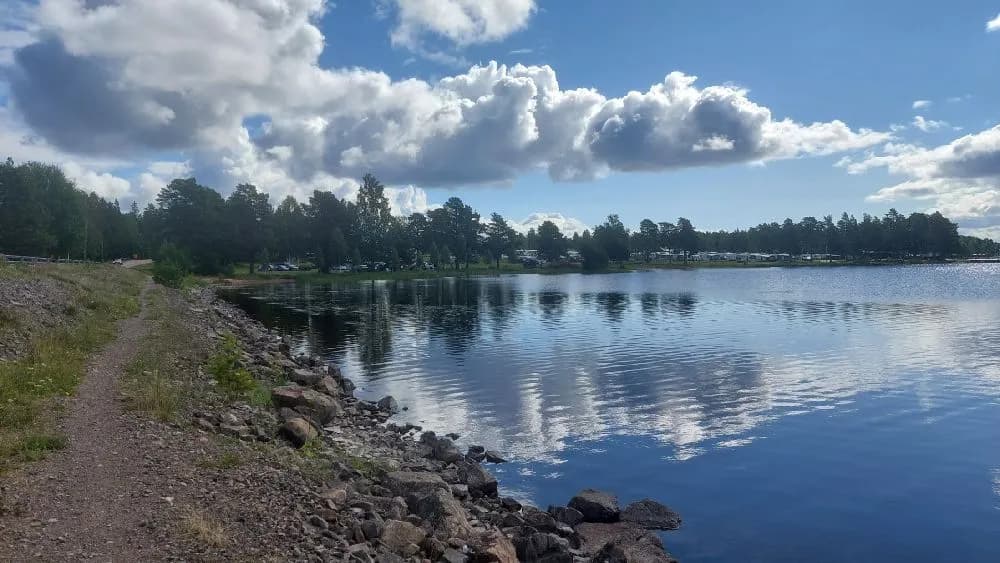 En klarblå himmel speglas i det lugna vattnet vid Sollerö camping, med omgivande grönska och några vita moln på himlen.