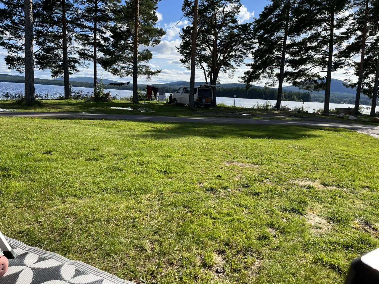 En grön campingplats med gräsmatta och skuggiga områden nära en kust eller en vik, omgiven av skog och belägen vid en insjö.