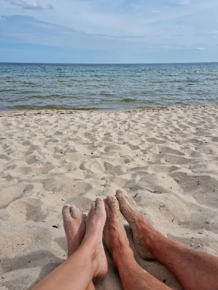 En person går barfota på en sandstrand vid kusten en solig sommardag. Havet syns i bakgrunden, vilket ger en känsla av semester och avkoppling.