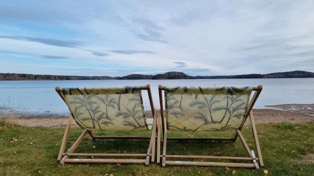 En vik med flera hopfällbara stolar i naturligt material står placerade på en sandstrand vid vattnet, omgivna av skuggor från träd.