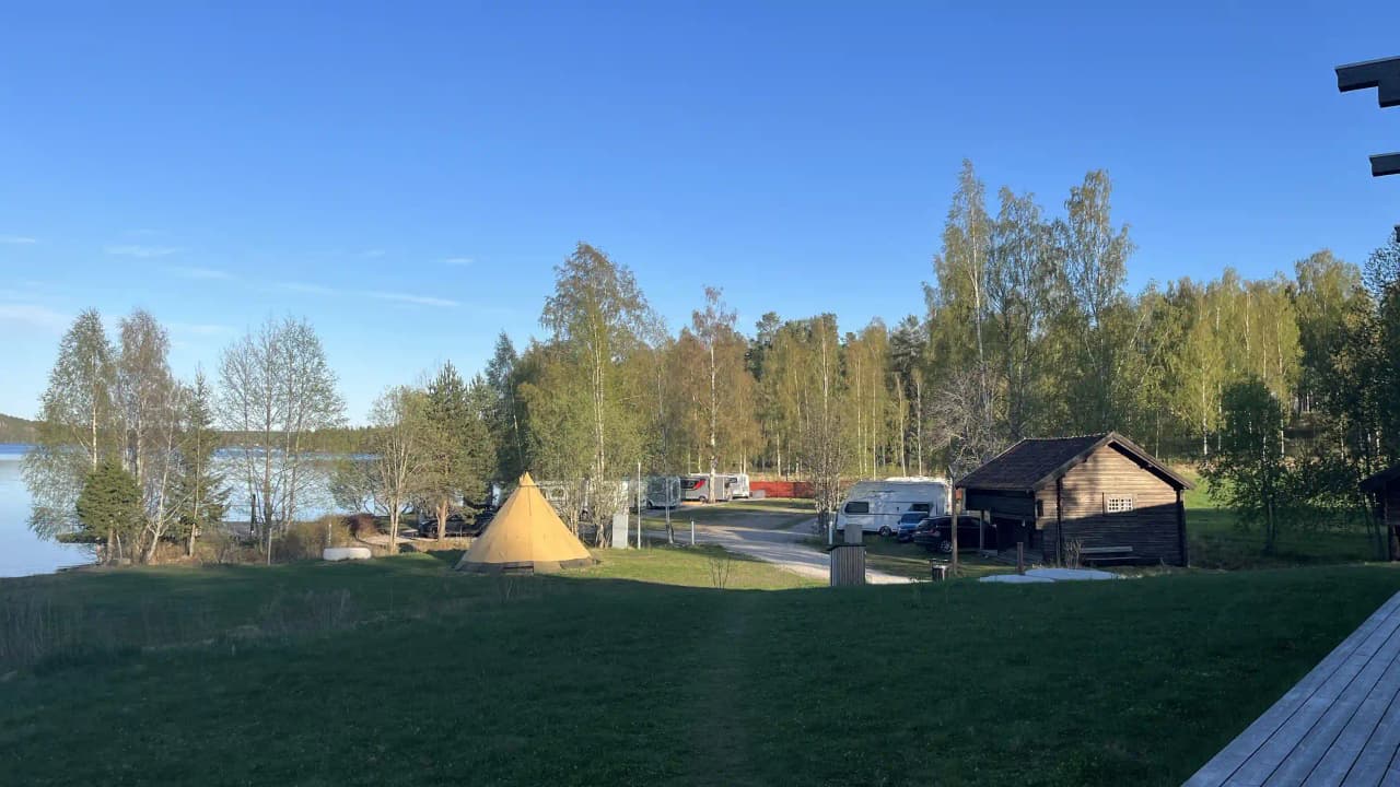 En stillsam plats med en liten stuga vid en sjö omgiven av lärkträd. En tältplats erbjuder campingmöjligheter i skuggan av träden.