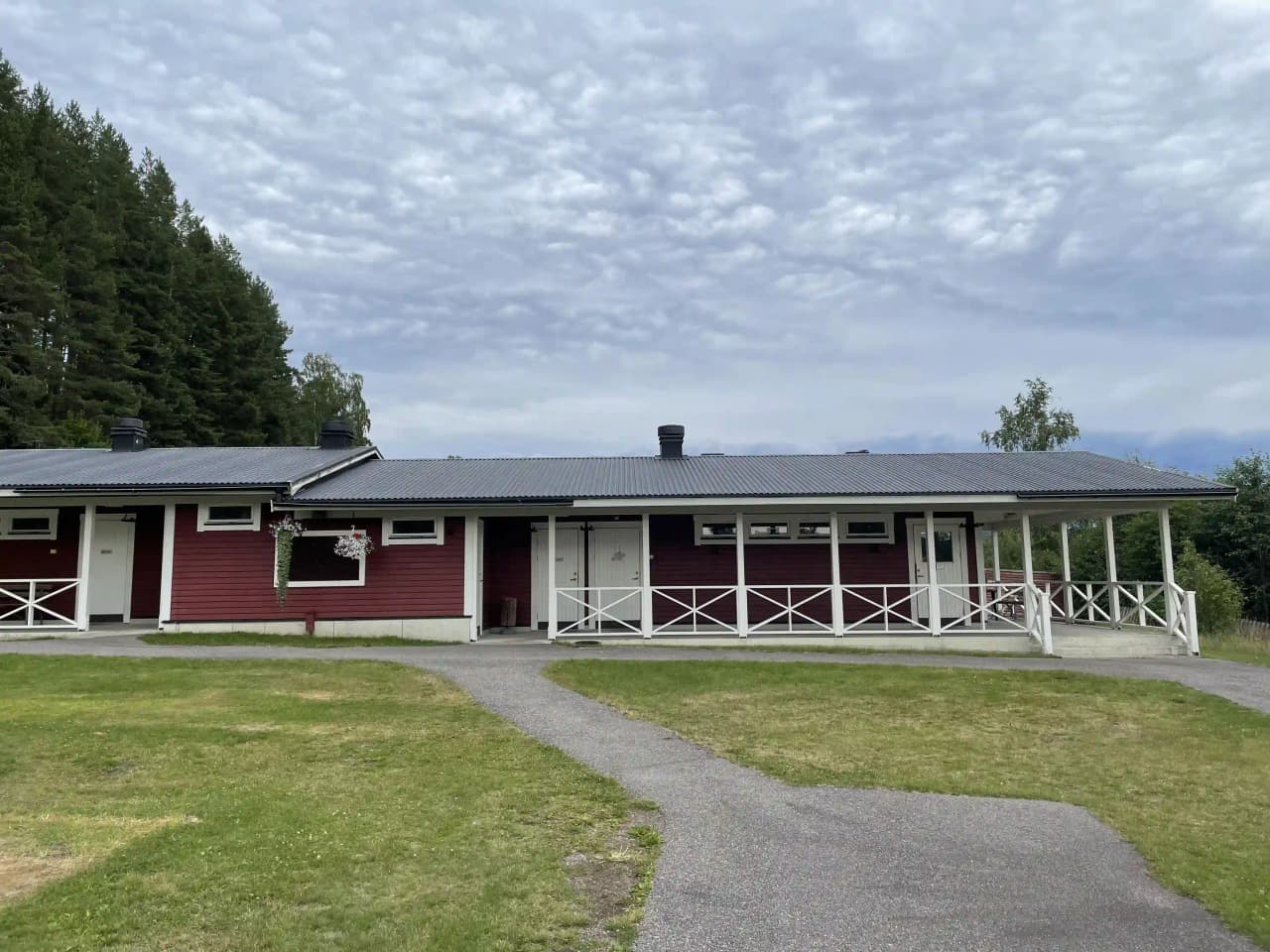 En liten röd stuga med vita knutar och veranda, belägen på en grön gräsmatta med omgivande träd och öppna ytor på Kävsta camping.