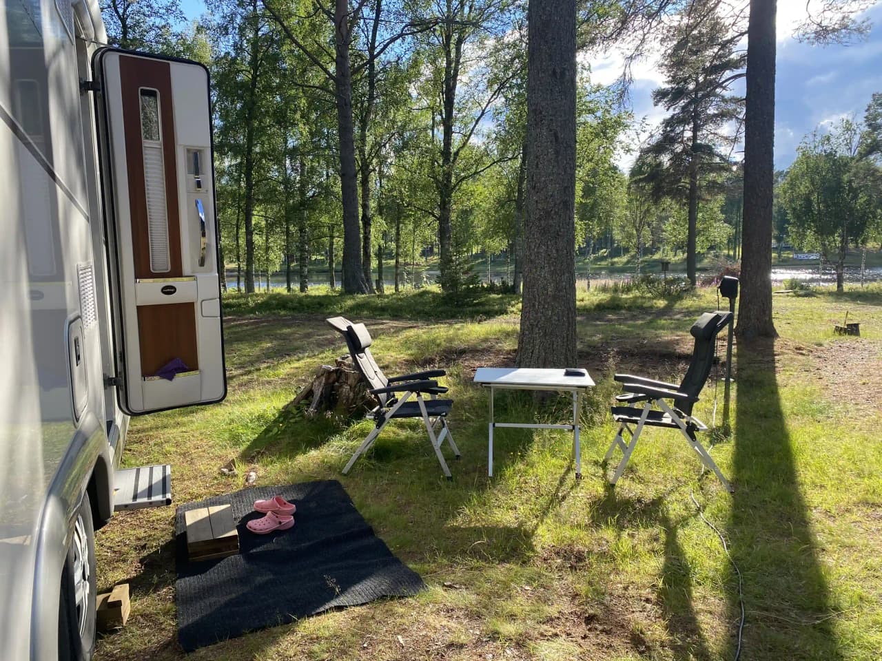 En campingplats med flera husvagnar och en husbil uppställda på en grön gräsmatta. I bakgrunden syns en skogsdunge och en cykel står lutad mot en husvagn. Solens skuggor faller över området och skapar en lugn atmosfär.