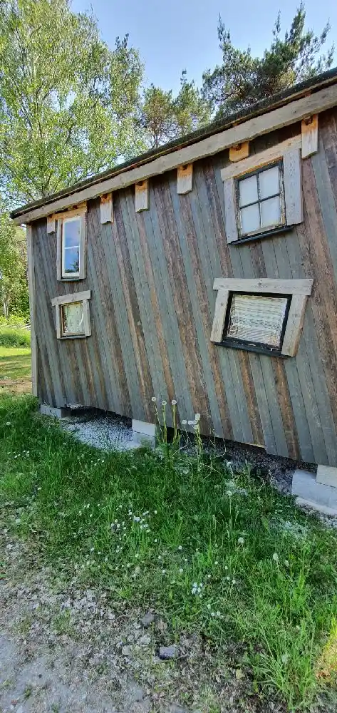 En trästuga i Fide Äventyrsby & Camping omgiven av grönska och blå himmel. Stugan har sidoklädd panel och en rustik känsla, vilket ger en inbjudande atmosfär för besökare som söker äventyr och avkoppling bland naturen.
