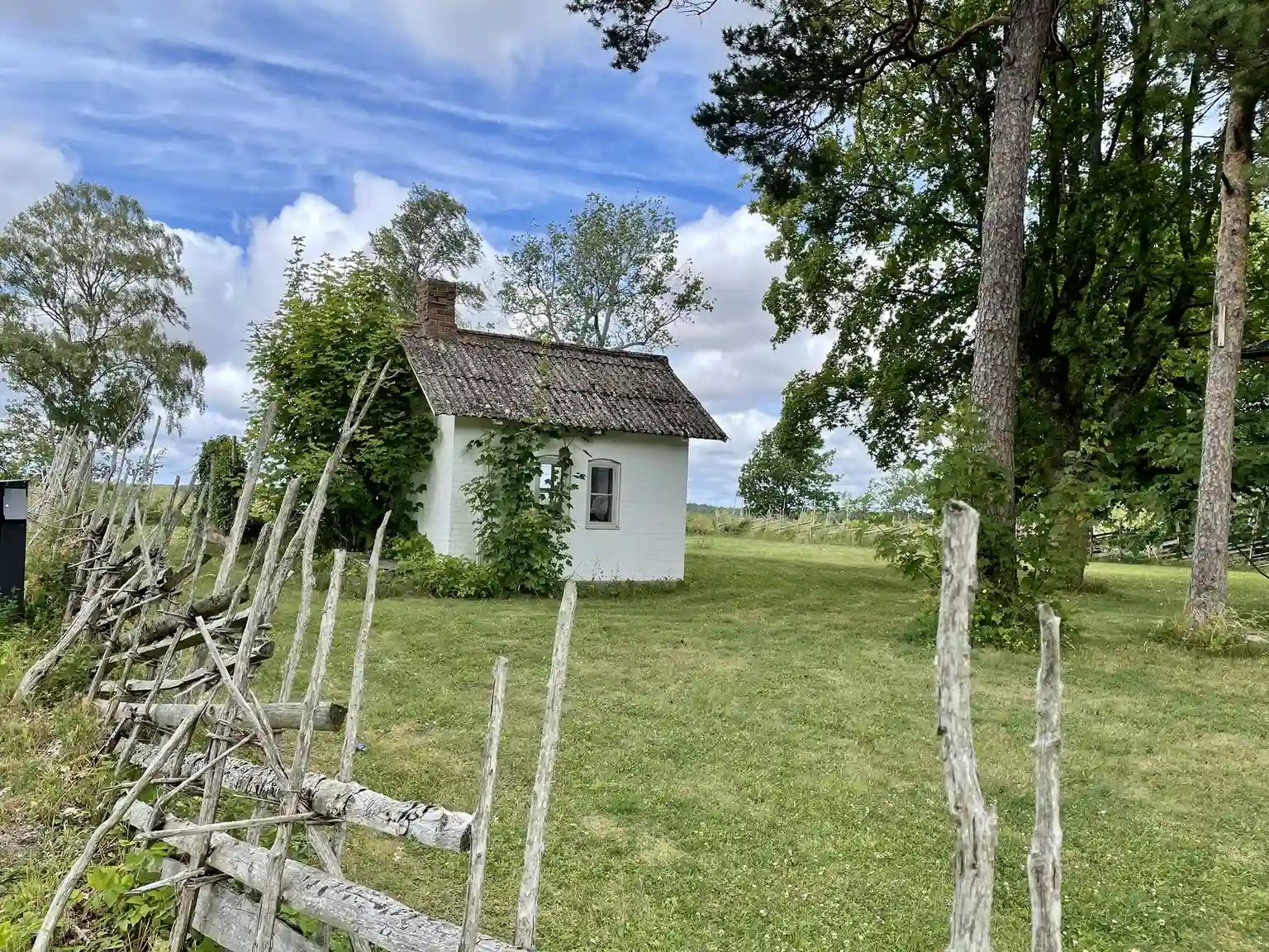 En idyllisk landskapsbild av Fide Äventyrsby & Camping, som visar en mysig stuga omgiven av grönskande buskar och gräsmattor, belägen i ett lantligt område.