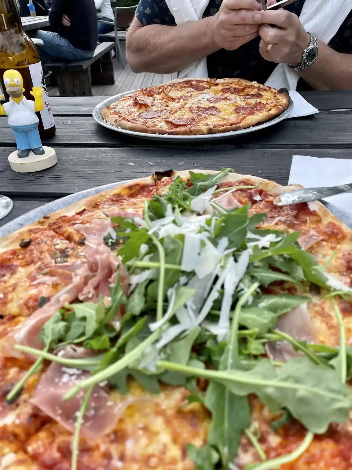 En närbild av en nygräddad pizza med olika köttingredienser, serverad på en tallrik på ett bord, omgiven av diverse köksredskap på Fide Äventyrsby & Camping.