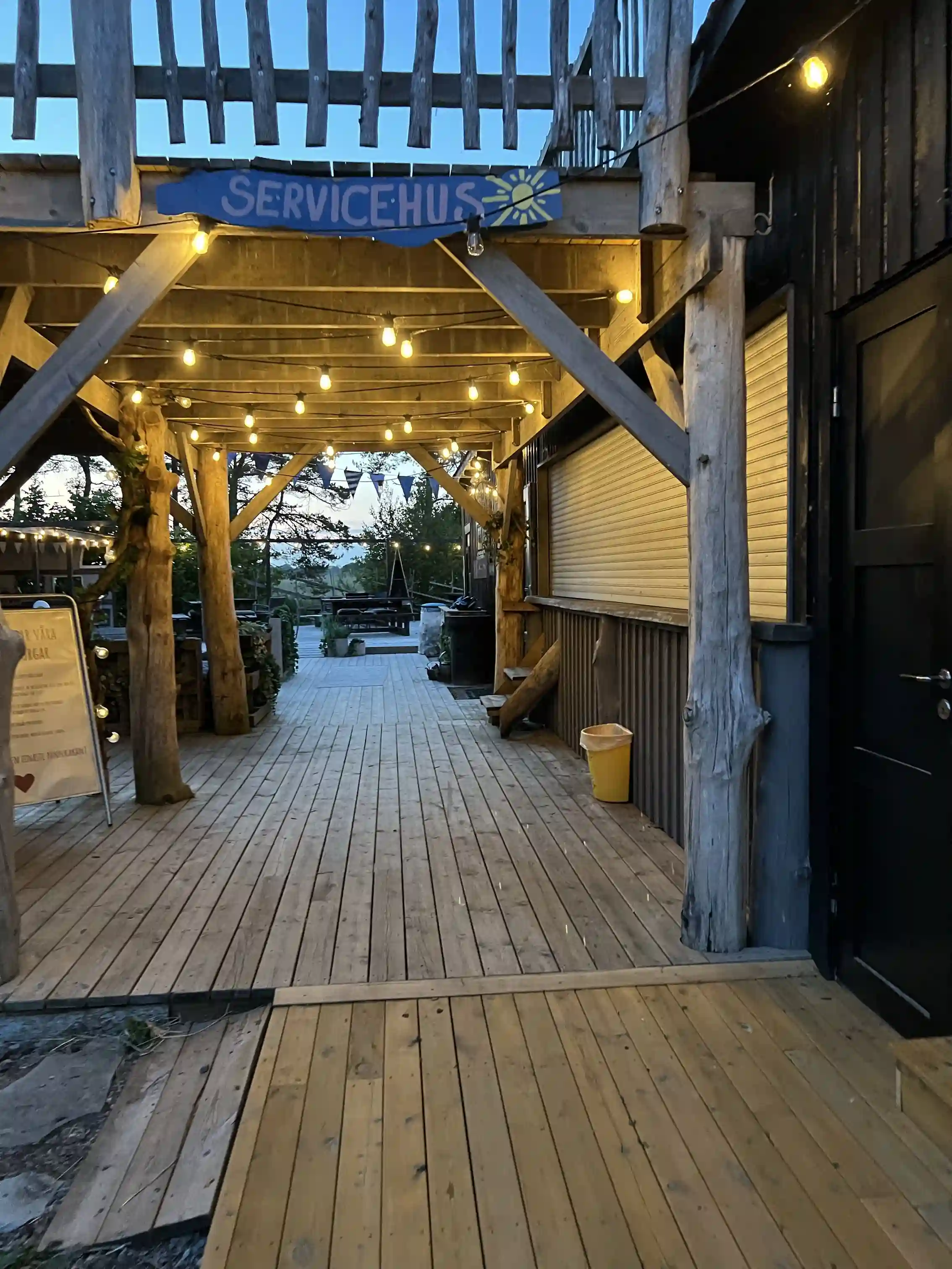 En träbyggnad med en veranda av träplankor vid Fide Äventyrsby & Camping. Verandan leder upp till en öppen entré med trappa. Naturmaterial såsom trä dominerar strukturen för en rustik och naturlig känsla.