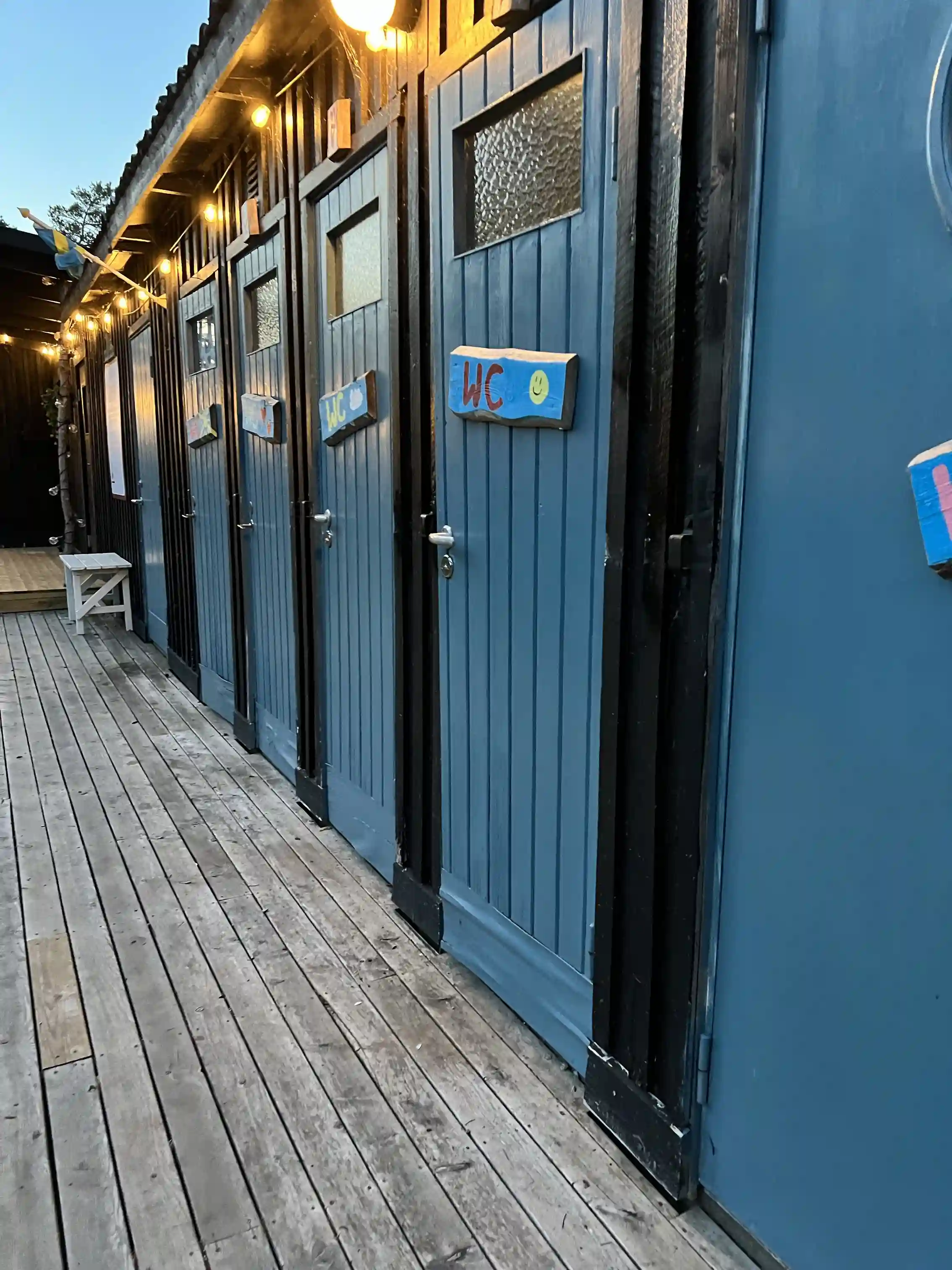 En träbrygga med metallräcke leder genom en naturskön miljö med träplankor och stålkomponenter, omgiven av frodig vegetation, vid Fide äventyrsby & camping.