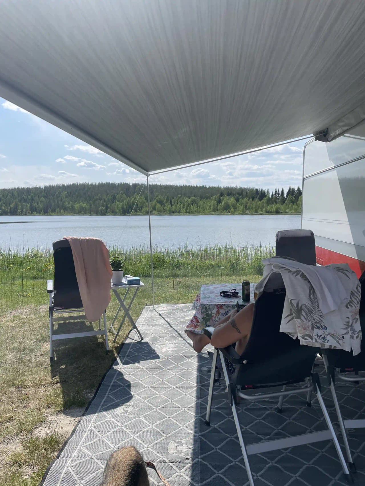 En campingplats med utemöbler under ett solskydd. I förgrunden finns ett bord och stolar, och i bakgrunden syns tält och tältöverdrag. Det är en solig dag och platsen ser inbjudande ut för avslappning och fritidsaktiviteter.