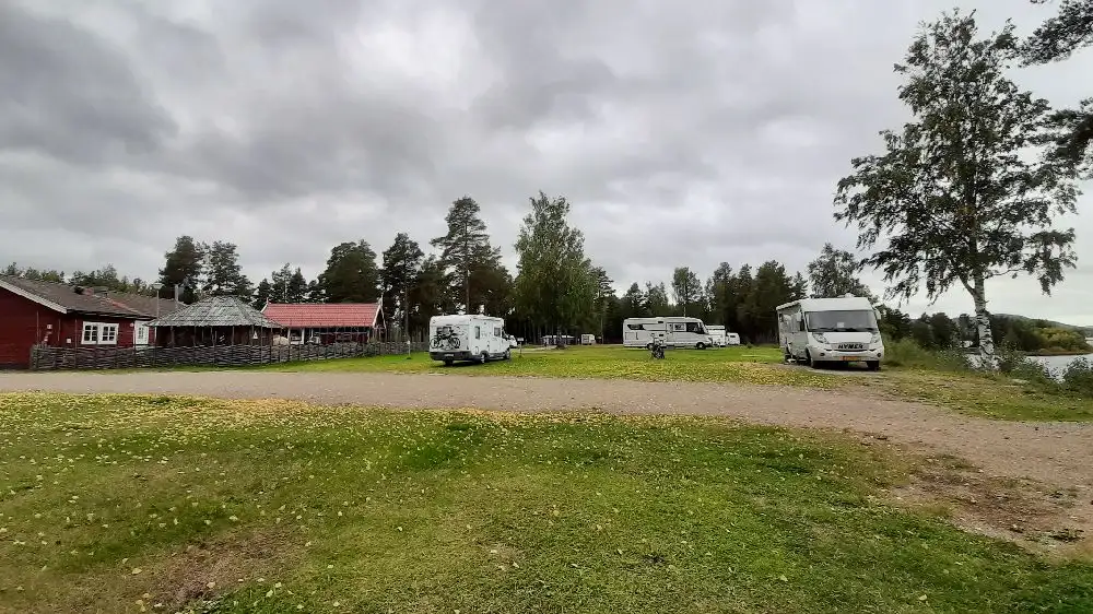 En bild av Ljusdals camping visar en parkeringsplats med flera fordon, inklusive en SUV, husbil och småbil. Tält är uppsatta i bakgrunden under en molnig himmel.