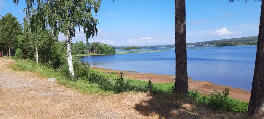 Utsikt över Ljusdals Camping med en blå himmel och vattendrag i förgrunden, omgivet av naturlandskap och horisont i bakgrunden.
