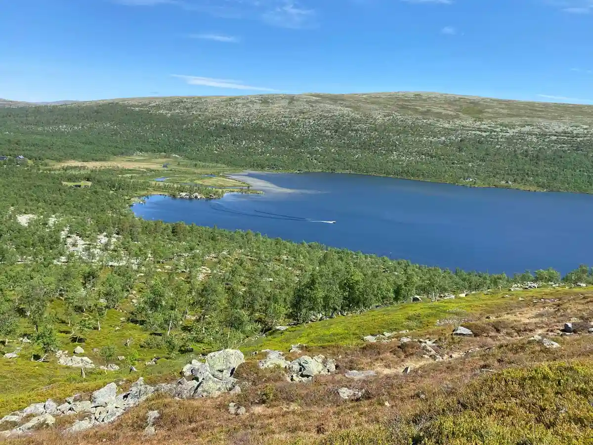 En pittoresk sjö omgiven av frodig natur och grässlätt, med klart vatten som reflekterar himlen. I bakgrunden finns en skogsbeklädd höjd, vilket ger en känsla av avskild vildmark.