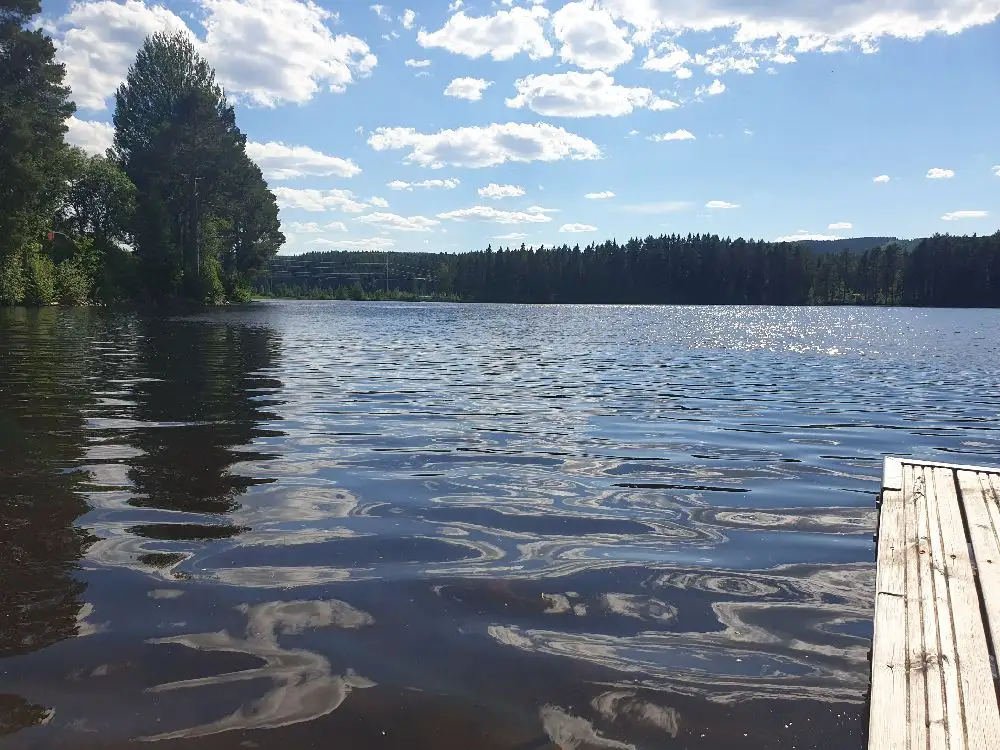 En sjö med klart blått vatten omgiven av grönskande stränder under en himmel med moln, typiskt för naturen kring Hotings camping.