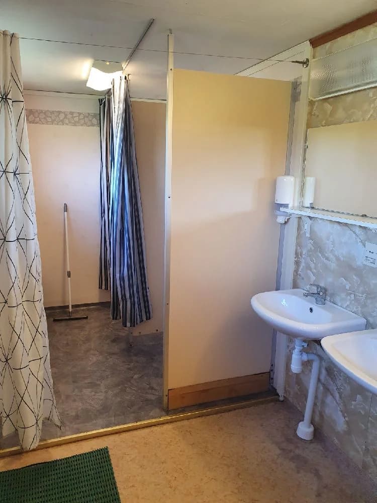 En ljus och välkomnande campingstuga på Hotings Camping, med ett stilrent inrett badrum. Badrummet har ljusa väggar, ett ljust klinkergolv och en dusch med duschdraperi. Det finns en modern sanitetsinredning och en inredning som är både funktionell och estetiskt tilltalande.