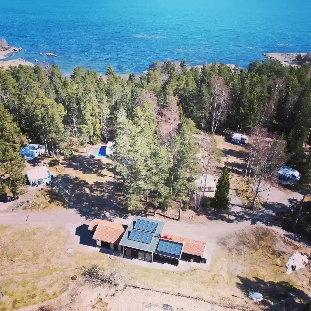 Flygbild av ängskärs havscamping som visar en naturskön kustlinje med träd, enkla stugor nära strandkanten och en lugn vik i bakgrunden.