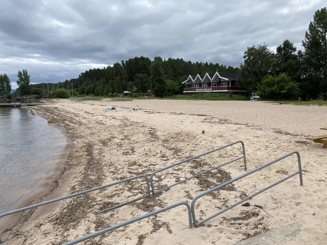 En sandstrand vid kusten med skuggiga områden från träd och växtlighet, omgivet av tropisk natur och en vik i bakgrunden.