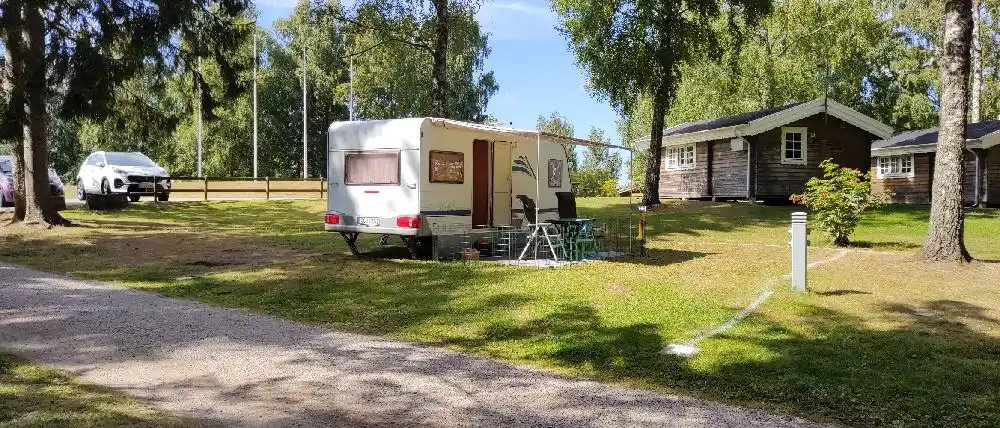 En somrig campingmiljö med stugor, husvagn och grönskande växter på Orebackens camping. Gräsmatta och parkerad husbil i en lantlig omgivning.