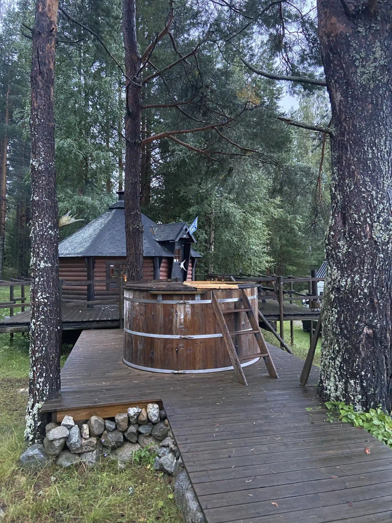 En timmerstuga i en skogsmiljö med trädkronor som ger skugga. Stugan har en utomhusstruktur med ett trädäck, belägen i en lummig och naturlig omgivning.