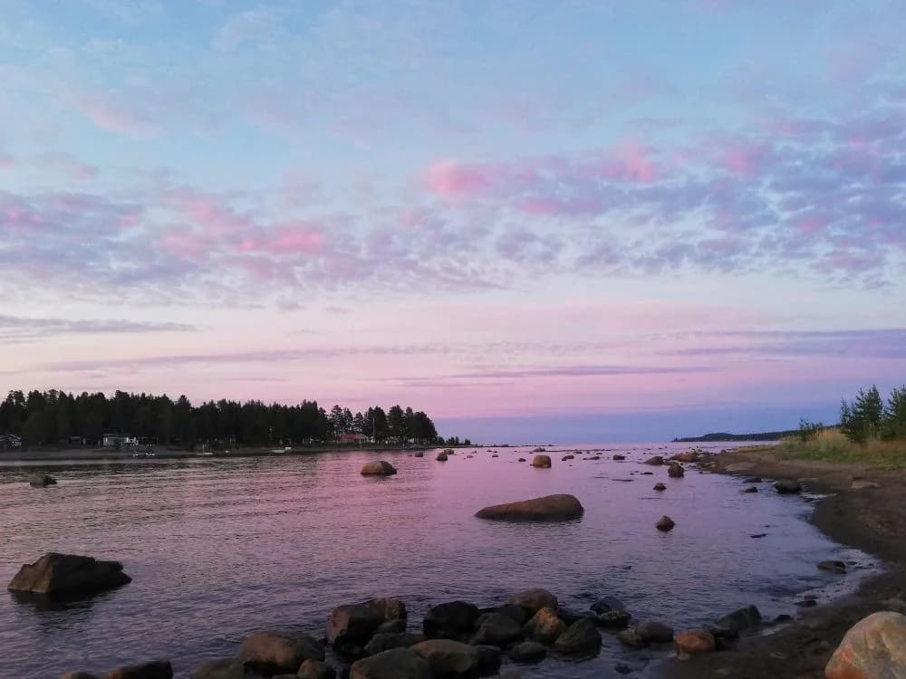 En lugn havsutsikt vid Bovikens havsbad i skymningsljus, med en klar horisont, stilla vatten och några klippor i förgrunden under en färgglad kvällshimmel fylld av moln.