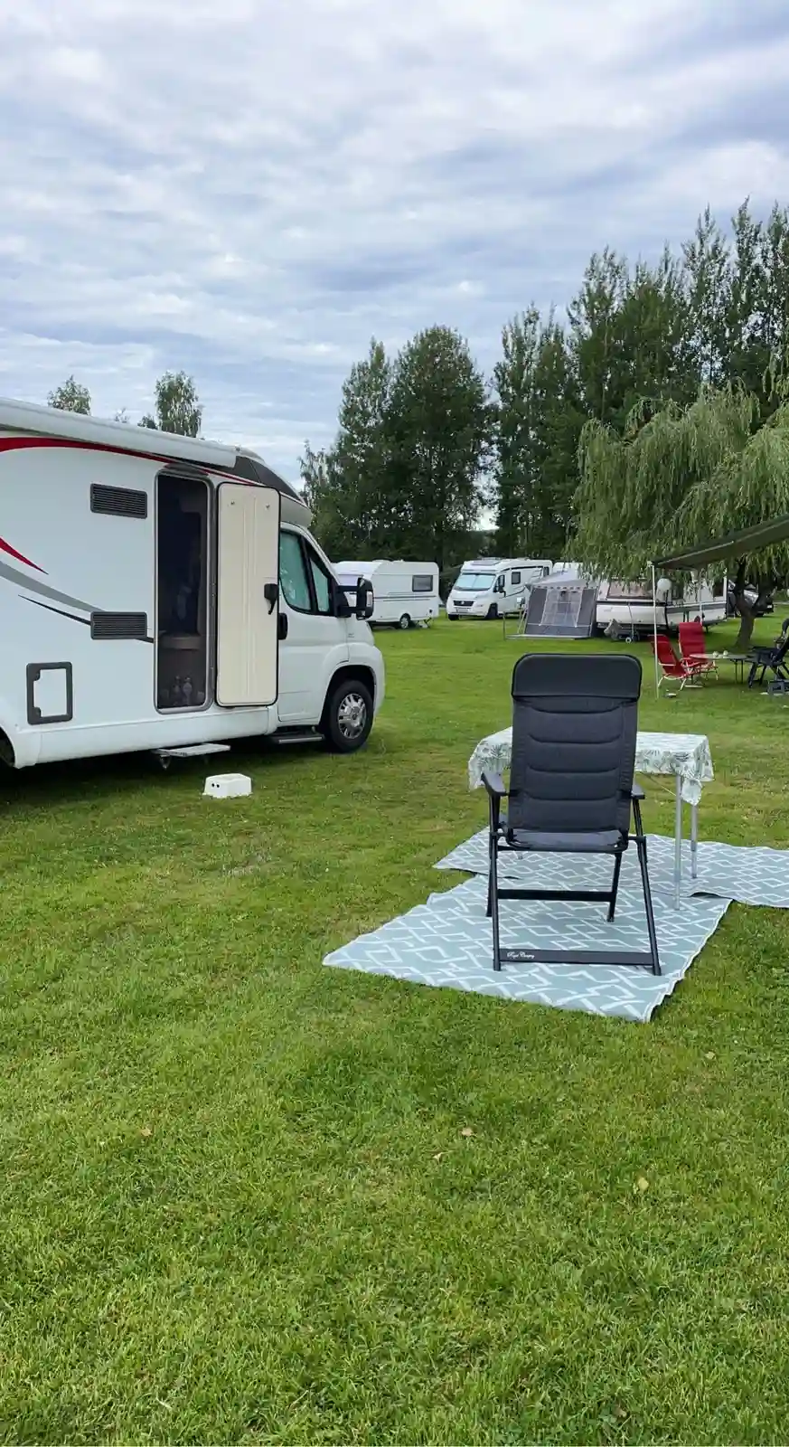 En husvagn parkerad på en grön gräsmatta med andra campingfordon i bakgrunden vid Säters camping. Omkringliggande träd ger skugga på campingområdet.