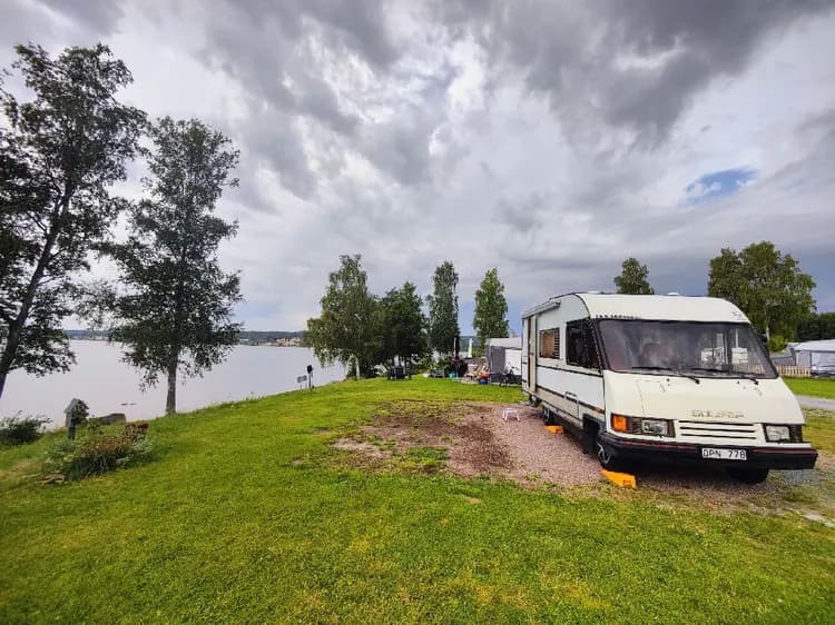 Bild av Säters camping med husvagnar och husbilar parkerade nära en sjö. Himlen är molnig med cumulusmoln och omgivningen är lantlig och lugn med grönska.