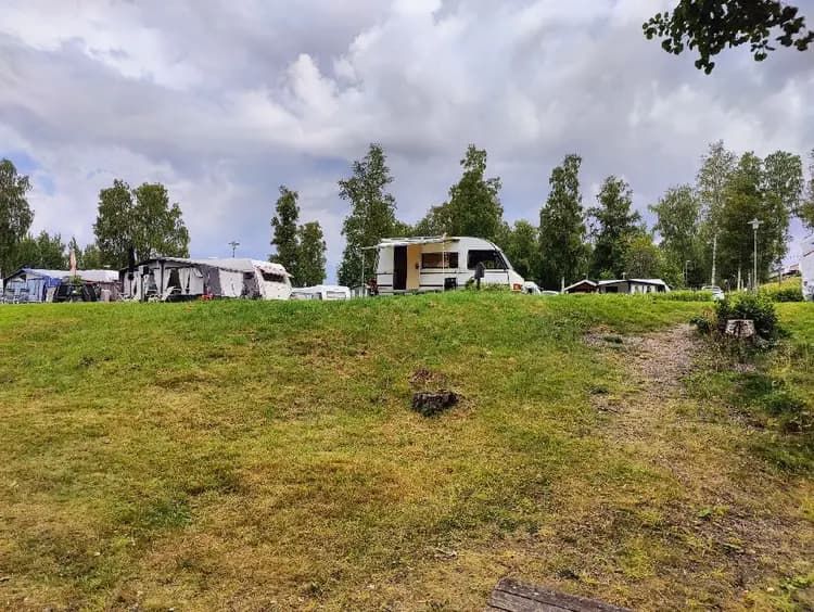En grön campingplats med husbilar och husvagnar uppställda på en äng. Mulna moln på himlen och omgivande grönska kompletterar det natursköna landskapet vid Säters camping.