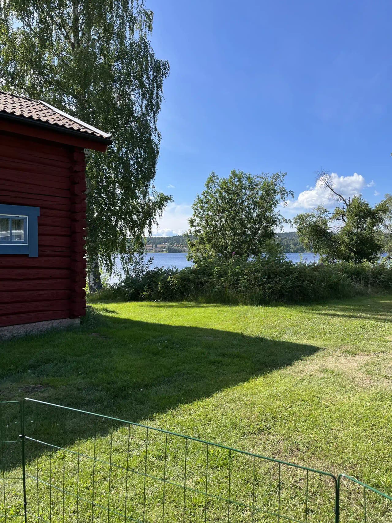 Bild av Säters camping: En grön campingplats med välskött gräsmatta, omgiven av staket och landskap. I bakgrunden syns träd och en vattenreservoar som ger en lugn och naturnära miljö.