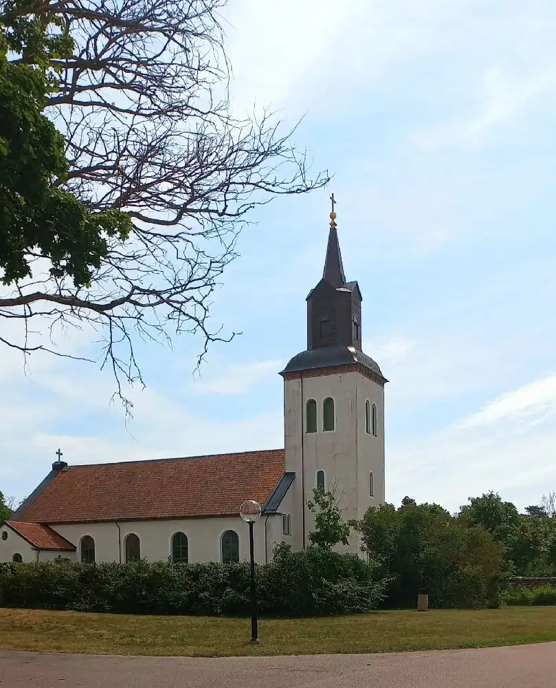 En vit kyrka med ett spetsigt torn reser sig mot en klarblå himmel omgiven av grönska.