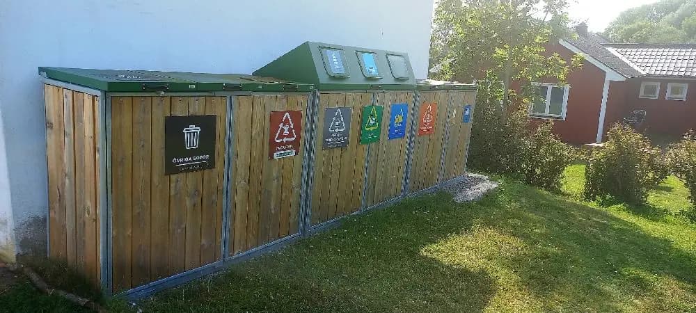 En återvinningsstation vid allégården i kastlösa, med träplankor och flera färgglada återvinningskärl för olika material.