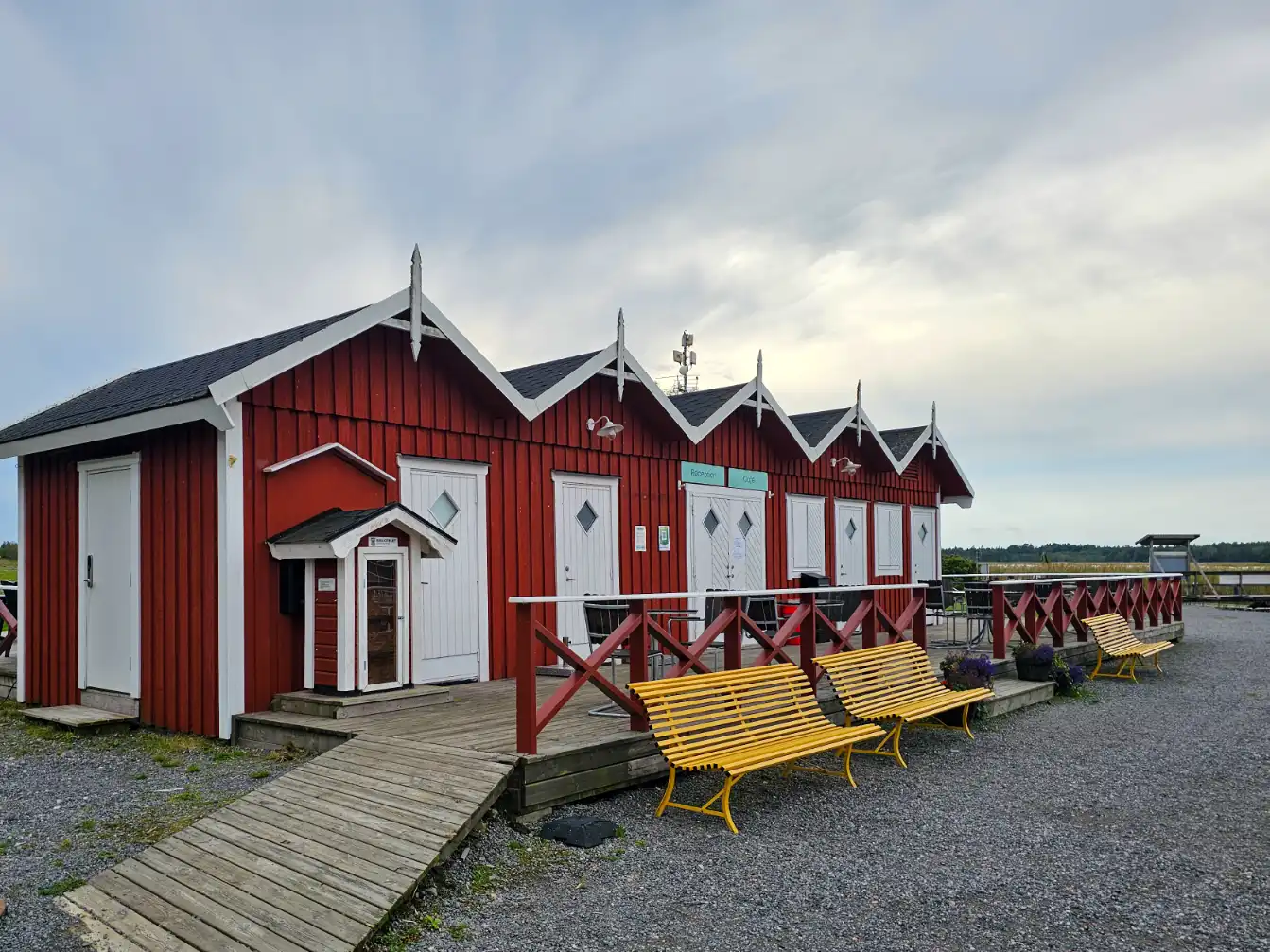 En träbyggnad vid vattnet med campingplats i bakgrunden, ljusa träpaneler och tak, del av Ekenäs gästhamn & camping.