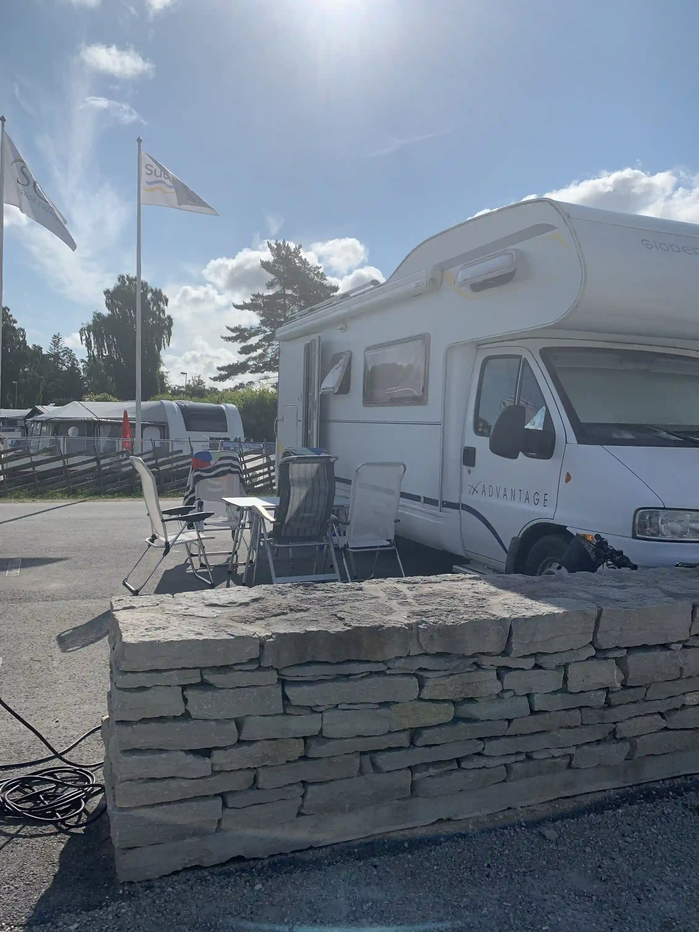 Bild som visar en parkeringsplats vid Sudersand Resort, med en stenmur i bakgrunden och flera parkerade fordon, inklusive skåpbilar och minivans, längs en uppfart med stenlagd mark.