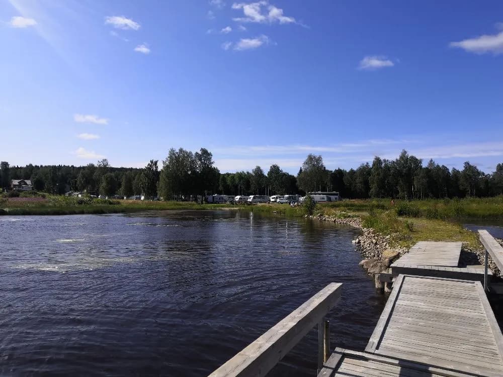 En naturskön campingplats vid en sjö med omgivande skog och vattenvägar. Solig sommardag med lummiga träd och klart vatten. Perfekt för friluftsliv och avkoppling.