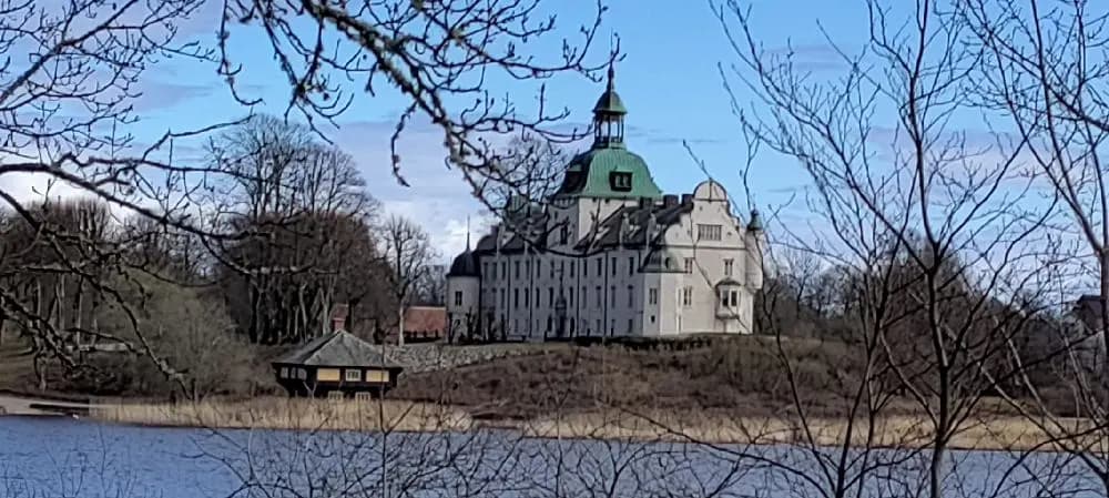 Bild av Stenrösets Camping i Trollhättan med utsikt över en vattenväg som omger en historisk byggnad med likheter till ett slott eller vattenslott. I bakgrunden syns ett vackert landskap med sjö och frodig grönska.