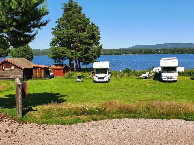 Bild av Västanviksbadets Camping i Leksand, visar en vacker kustlinje vid en sjö med ett campingområde där husbilar är parkerade. Sjön omges av frodig grönska och landskapet ger en fridfull och avkopplande atmosfär för besökarna.