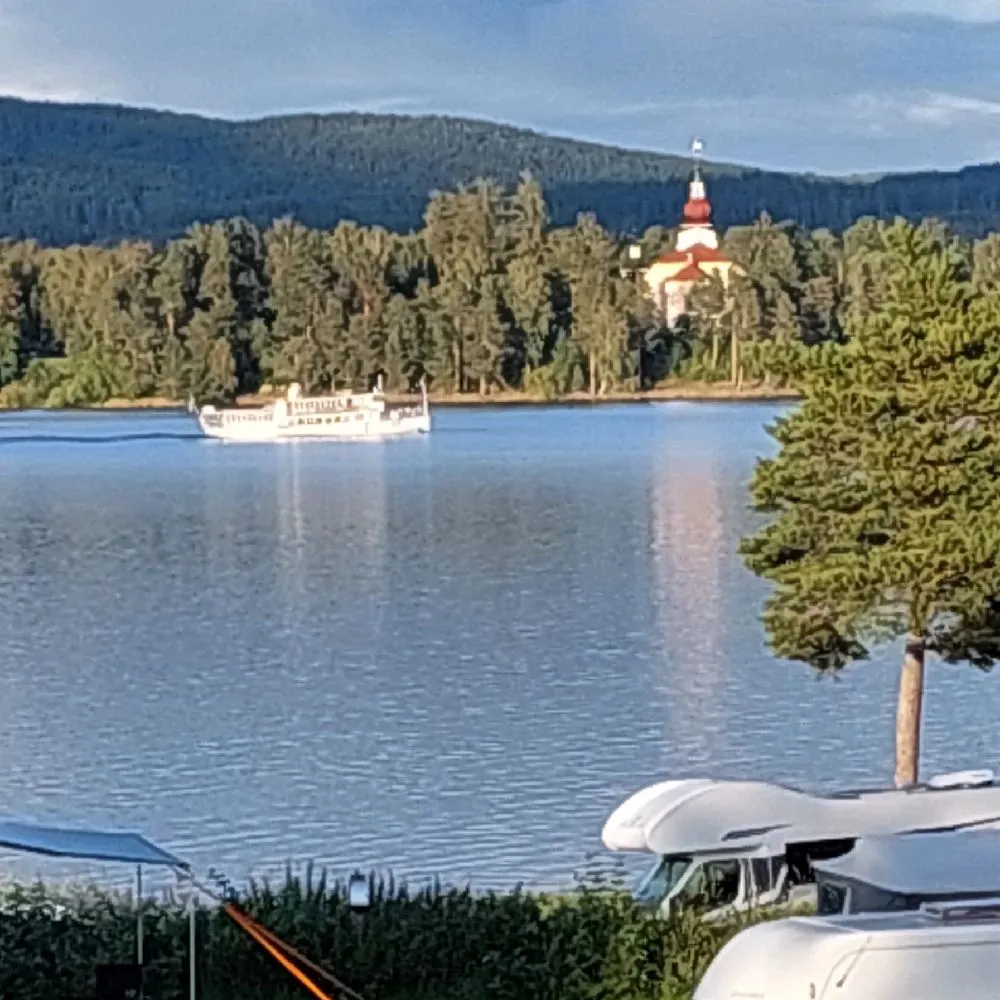 En vy över Västanviksbadets camping i Leksand med en sjö i förgrunden där en båt ligger vid den lugna vattenytan, omgiven av natursköna stränder och frodig grönska i bakgrunden.