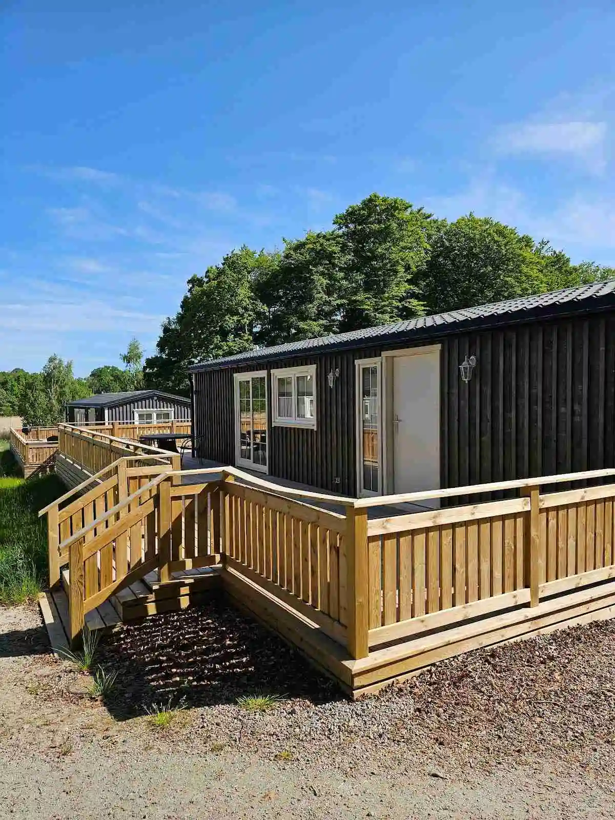 En mobil hem på en campingplats med ett trädäck i trä. Hemmet är omgärdat av ett staket och har sidobyggnader inplacerade i en prydlig trädgårdsmiljö.