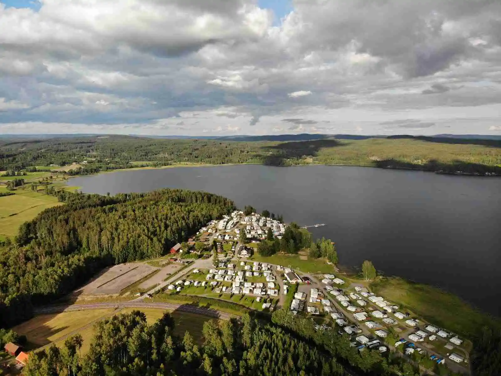 Flygbild över Haganäset vid Charlottenberg camping & stugor, med en sjö och kustlinje i förgrunden som omges av grönska och skogslandskap.