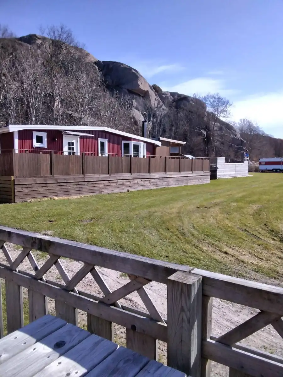 En mysig campingmiljö med en stuga omgiven av en grön gräsmatta. Det finns en uppfart och ett picknickbord med bänkar utomhus, vilket bjuder in till avkoppling i friluftslivet.