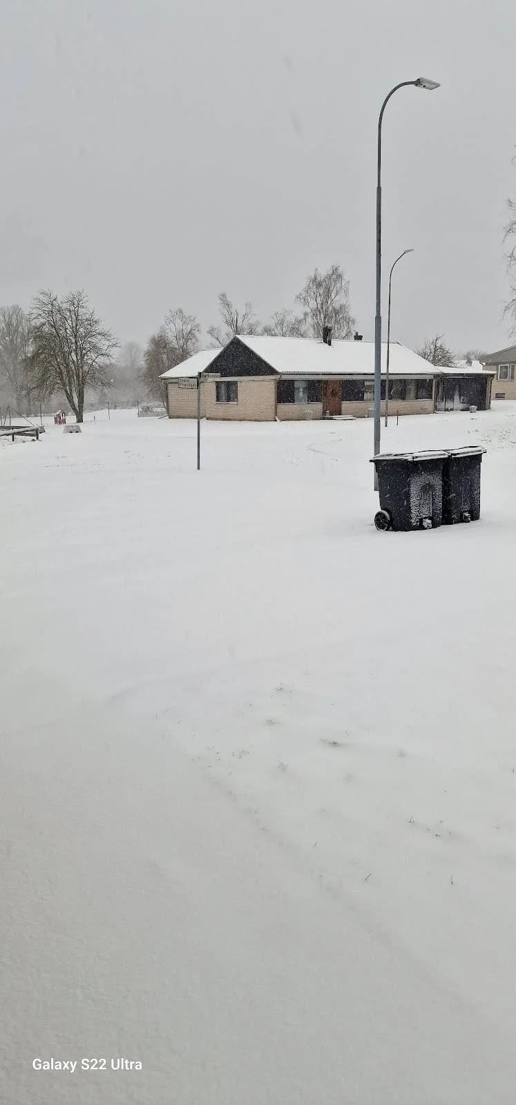 En snötäckt campingområde med ett litet skjul i bakgrunden, omgiven av vinterlandskap och pågående snöfall.