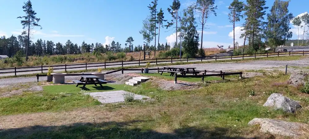 En bild av ett friluftsområde vid Gullviks Havsbad Camping & Stugby med picknickbord och bänkar. Moln syns på himlen och området omges av öppna fält och stigar.