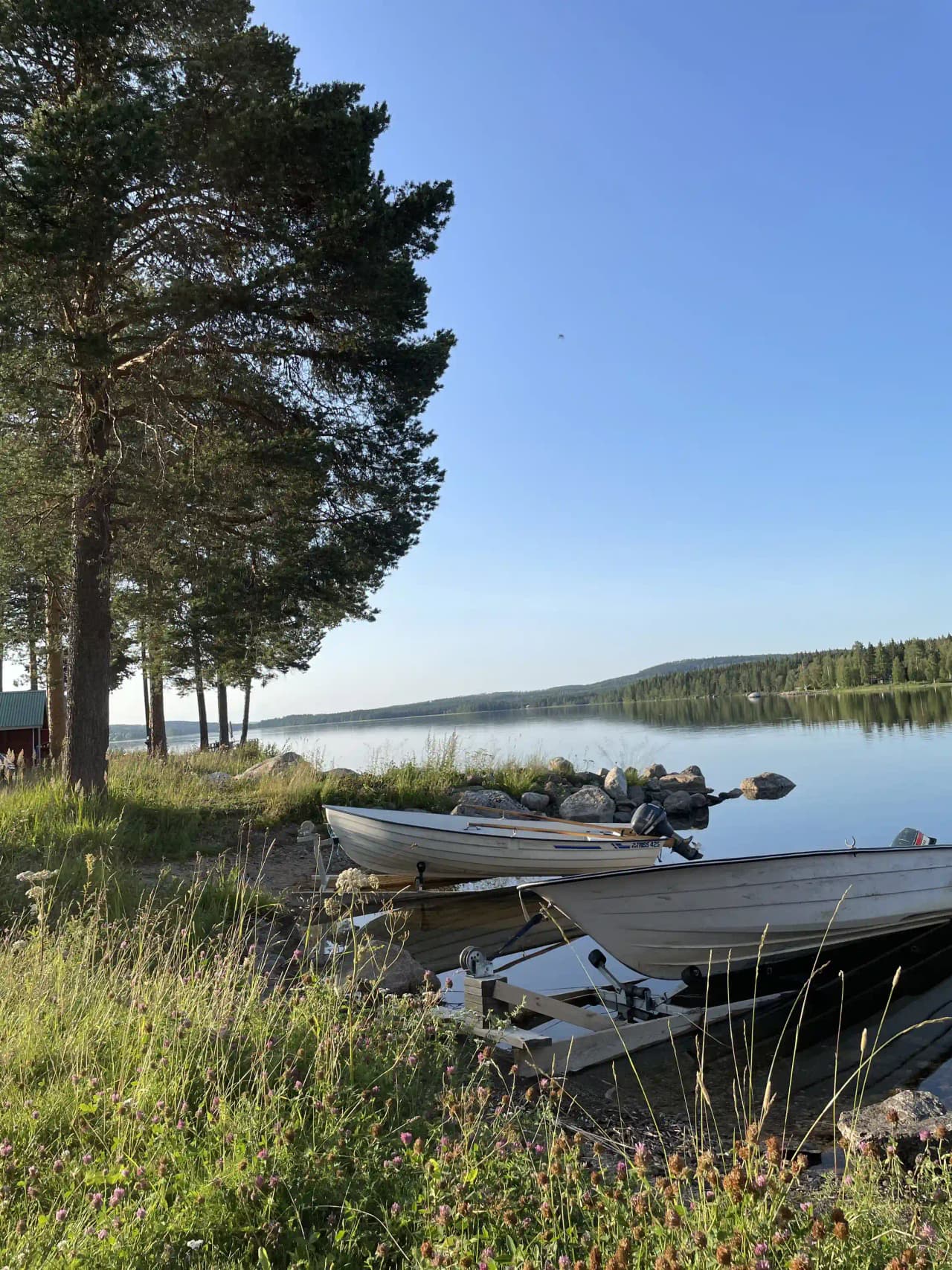 En naturskön vy över en vik med klarblå himmel och ett lugnt vattendrag där flera båtar ligger förtöjda längs stranden, omgivet av grönskande natur.
