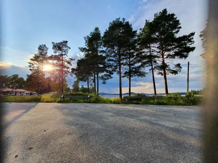 En asfalterad väg slingrar sig genom ett naturskönt landskap med grönskande träd och en klar himmel. Solsken bryter igenom molnen och kastar ljus över omgivningen.