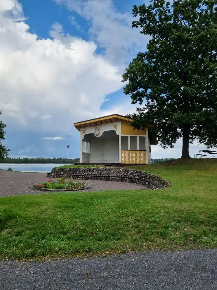 En grönskande park med en välskött gräsmatta. I mitten av bilden står en paviljong omgiven av träd och annan växtlighet. Det finns också en kiosk i bakgrunden. Perfekt för avkoppling och utomhusaktiviteter.