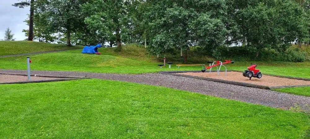 En grönskande park med välskötta gräsmattor, omgivna av träd och buskar. I förgrunden syns lekutrustning för utomhuslek, lämplig för campingbesökare som söker rekreation och avkoppling.