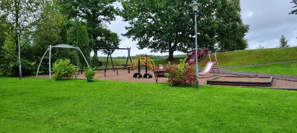 En grönskande campingplats med lekutrustning och rutschkana i en offentlig parkmiljö, omgiven av träd och gräs.