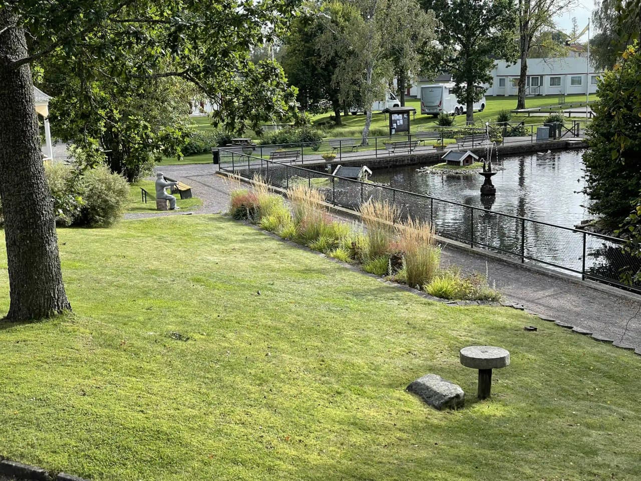 En grönskande park med frodig växtlighet och en liten damm. I förgrunden syns en träbänk med utsikt över den natursköna omgivningen vid sjöparkens camping.